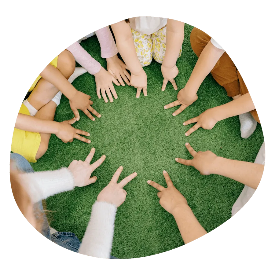 Un gruppo di bambini seduti in cerchio sull'erba con le mani al centro, durante un'attività CLIL del corso estivo Move Your Summer